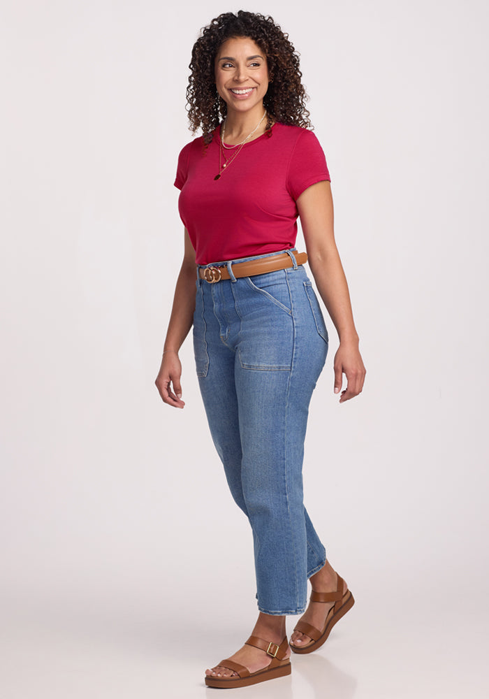 Woman with curly hair in a Woolx Addie Crew Neck (Poppy Spark), blue jeans, brown sandals, and a belt, smiling.