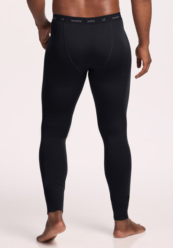 A man stands barefoot on a light background, seen from the back, wearing fitted black Woolx Backcountry Leggings, featuring a visible waistband labeled "woolx.