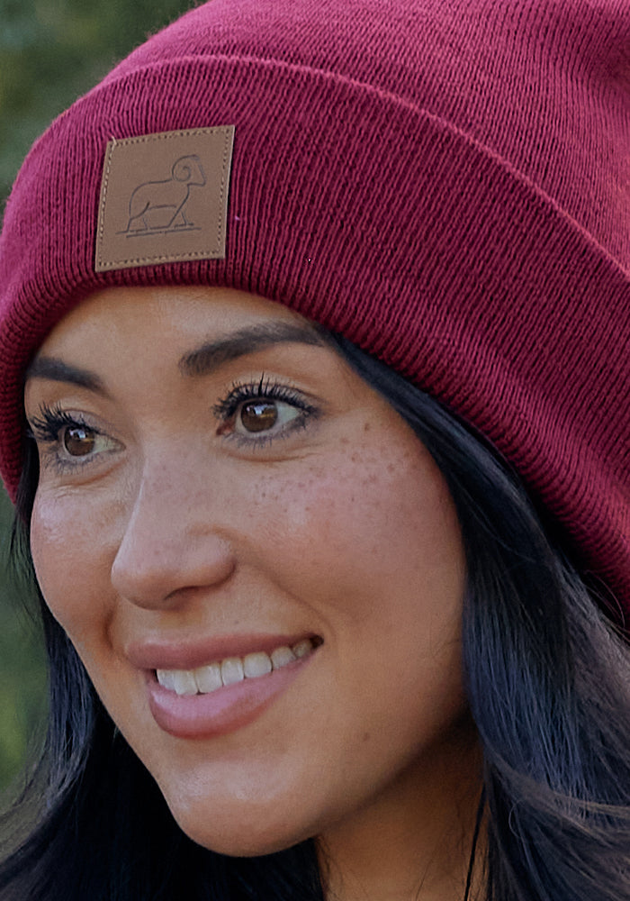 A woman with long dark hair smiles while wearing the Woolx Baylor Beanie in Toasted Garnet, featuring a brown patch with a simple ram illustration—a cozy merino wool winter hat for chilly days.