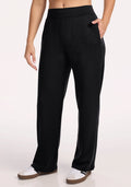 A person wears Woolx Bree Lounge Pants in black, hands in pockets, paired with white sneakers with brown soles, standing against a plain light background.