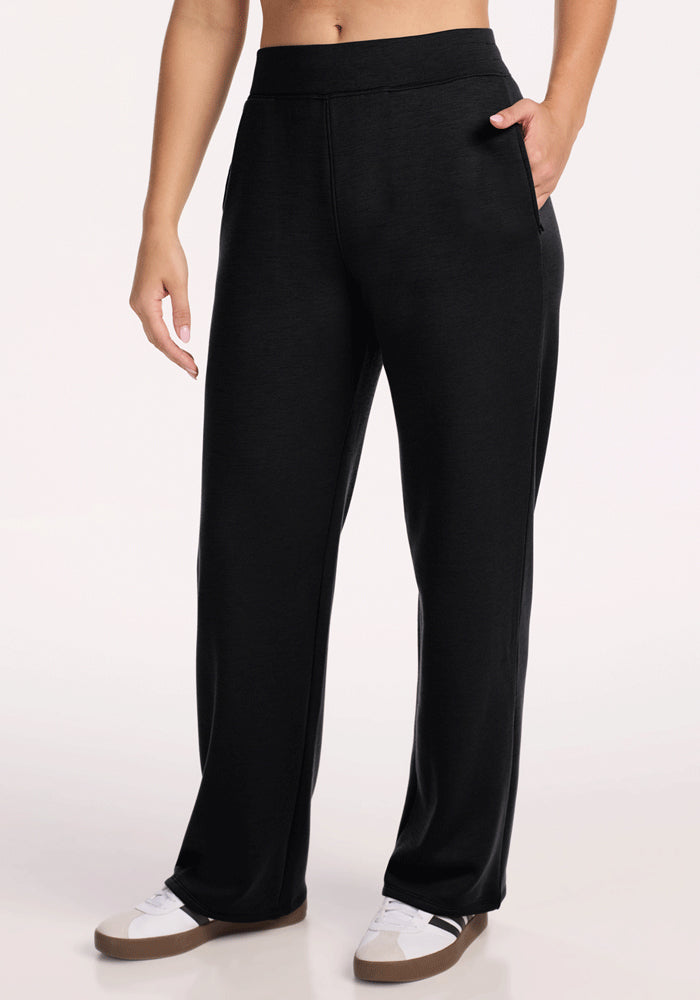 A person wears Woolx Bree Lounge Pants in black, hands in pockets, paired with white sneakers with brown soles, standing against a plain light background.