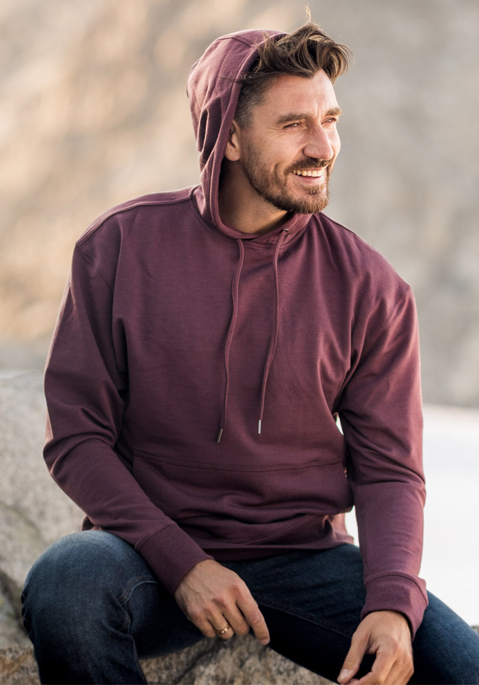 A bearded man smiles outdoors in a maroon Woolx Chase Hoodie • Final Sale - Grape Harvest and dark jeans, seated against a blurred natural background of sand or rocks.
