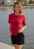 Woman in a Woolx Delia Ribbed Short Sleeve - Poppy Spark smiles by the waterfront, buildings behind her.