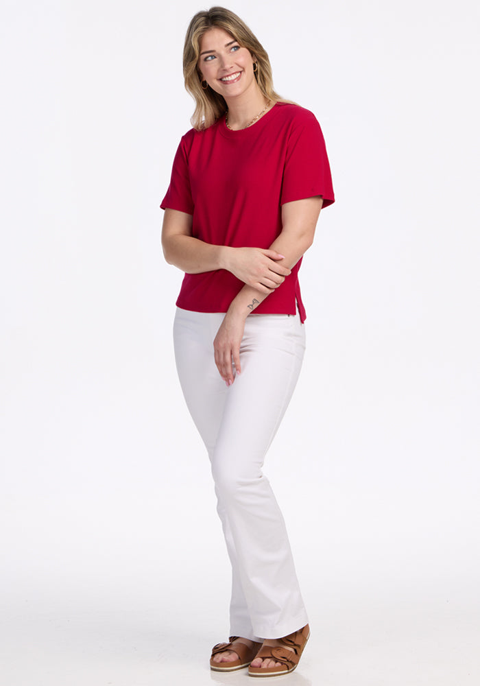 Smiling woman in Woolx Delia Ribbed Short Sleeve in Poppy Spark, white pants, brown sandals, on a white background.