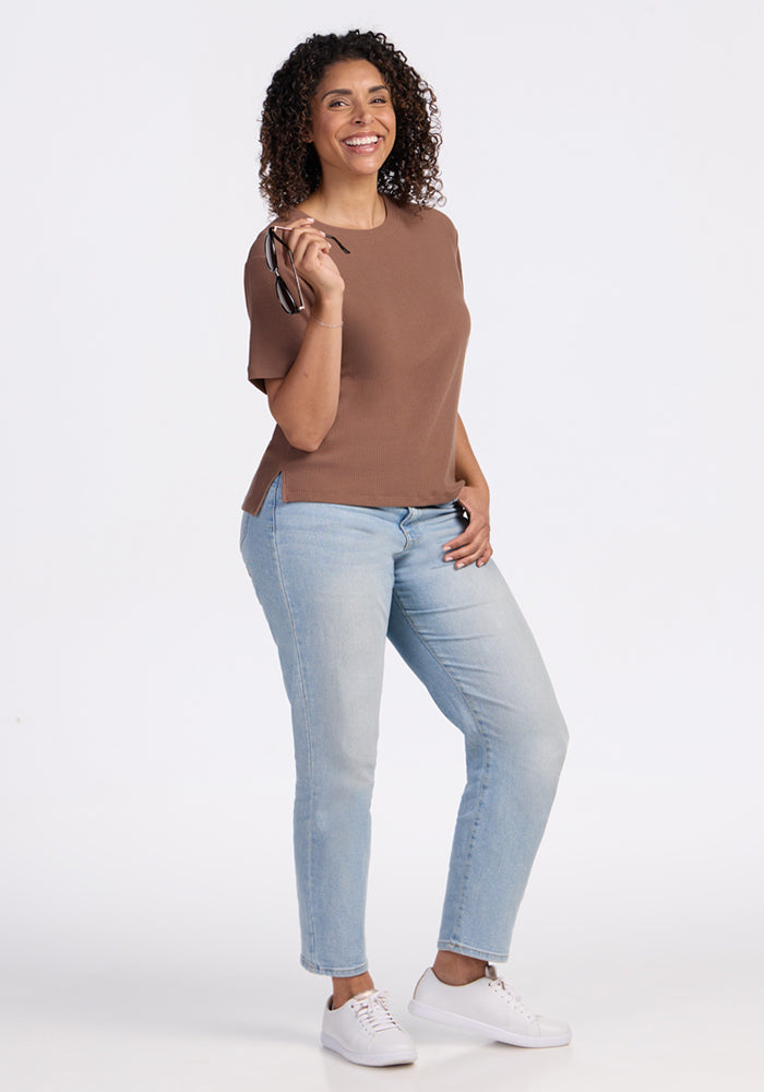 Smiling woman wears Woolx Delia Ribbed Short Sleeve in Sienna Kiss and light jeans, holding eyeglasses.