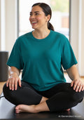 Smiling woman with dark hair wears Woolx Delia Ribbed Short Sleeve – Surfside, cross-legged on a mat indoors.