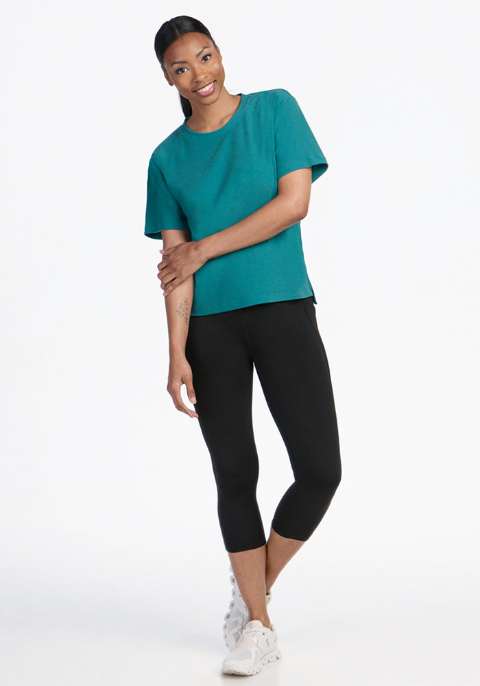 Woman smiling in the Woolx Delia Ribbed Short Sleeve in Surfside, black leggings, and white sneakers on white.