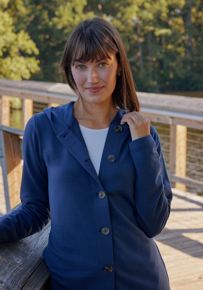 A woman with straight brown hair and bangs wears the Woolx Edna Hooded Cardigan in Cream Heather over a white top, standing on a wooden walkway by the water with trees and sunlight in the background.