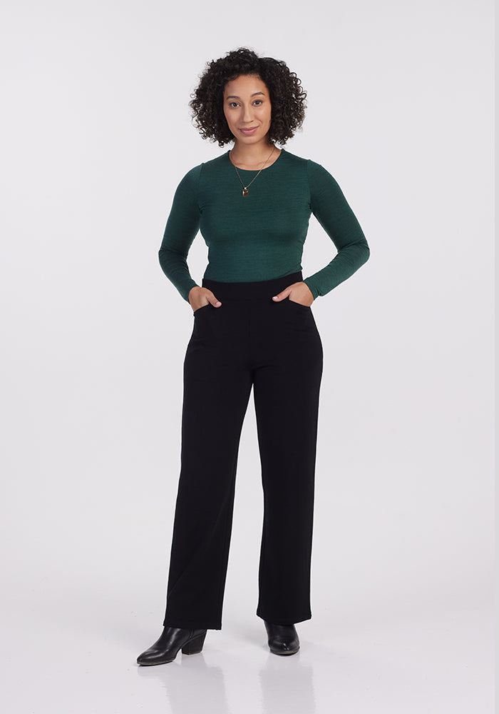 A person with curly hair stands against a neutral background, wearing a green long-sleeve shirt, Ellie Wide Leg Pants from Woolx, and black boots. Their hands rest casually in their pockets, exuding comfortable style and warmth.