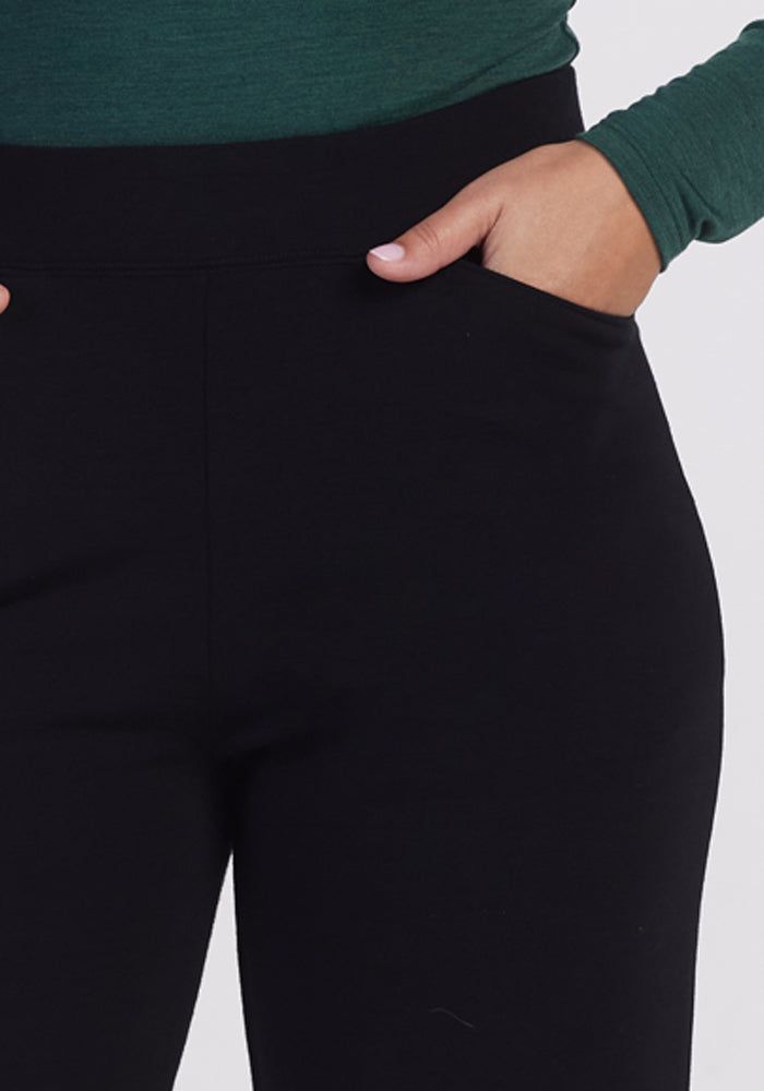 A person wearing Woolx's Ellie Wide Leg Pants in black and a green long-sleeve shirt is shown with a hand in one pocket of the pants. The image focuses on the waist and upper legs, highlighting the warmth and comfort of these premium fabric pants.