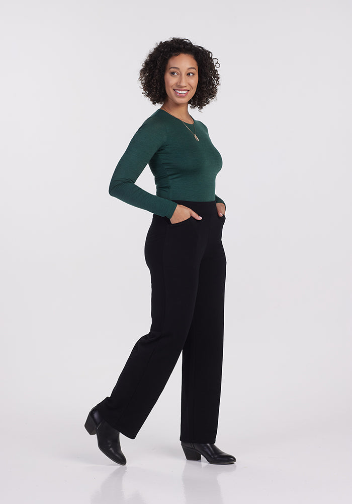A woman with curly hair smiles while wearing a green long-sleeve top and the cozy Ellie Wide Leg Pants by Woolx in black. She stands with her hands in her pockets, turning slightly to one side against a plain white background.