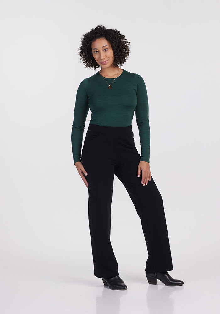 A person with curly hair stands against a plain background, wearing a green long-sleeve top, Woolx Ellie Wide Leg Pants in black, and black boots. They are posing with one hand on their hip and the other relaxed by their side.