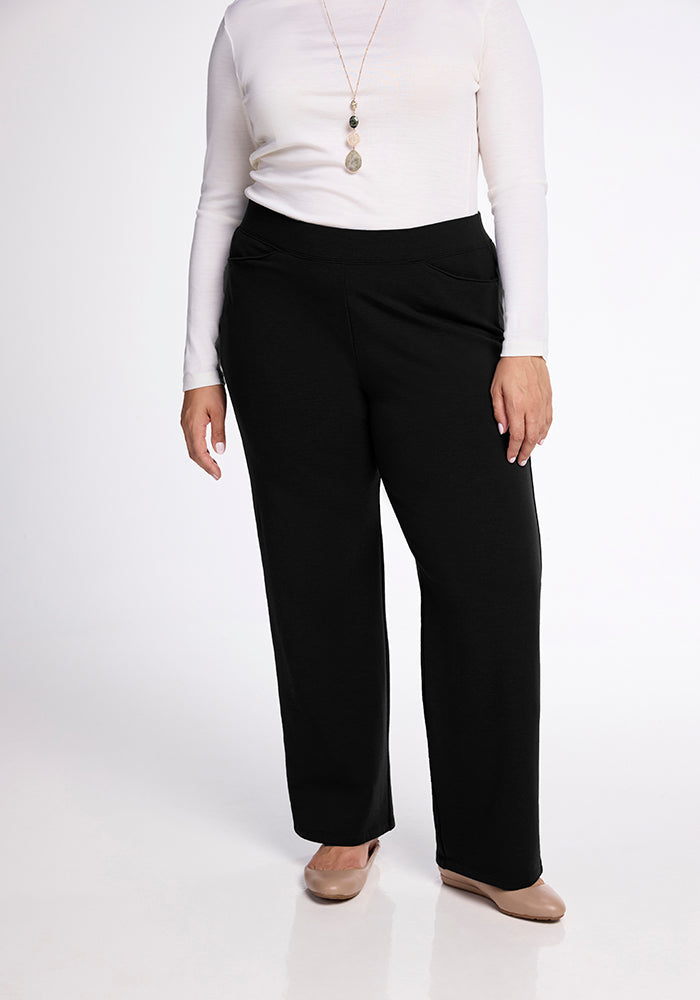 A person wearing a long-sleeved white top, beige shoes, and Woolx Ellie Wide Leg Pants in black. These cozy pants pair perfectly with their long necklace with pendants. The background is plain white.