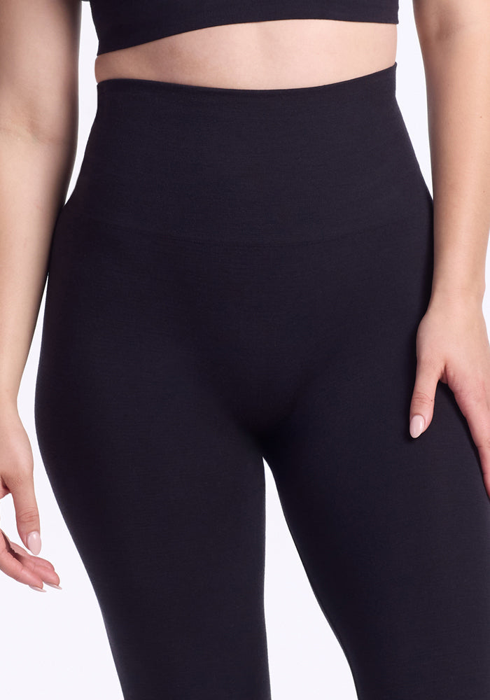 A person wears Woolx Ellis Seamless Petite Tights in black, featuring tummy control, paired with a black crop top. Shown from midriff to thighs with one hand by their side against a plain white background.
