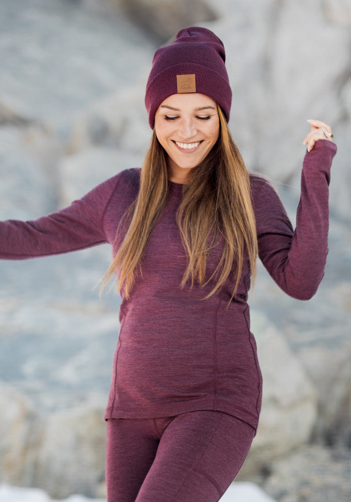 A woman wearing the Woolx Elsa Long Sleeve • Final Sale - Grape Harvest Melange smiles outdoors in a snowy, rocky landscape, paired with a matching maroon beanie and leggings.