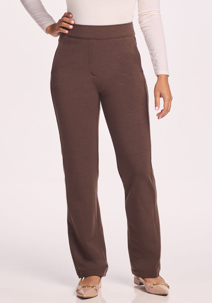 A person wearing Woolx Emerson Straight Leg Pants in Bracken, paired with a white long-sleeve top and beige flats, stands with one hand on their hip against a plain light background.