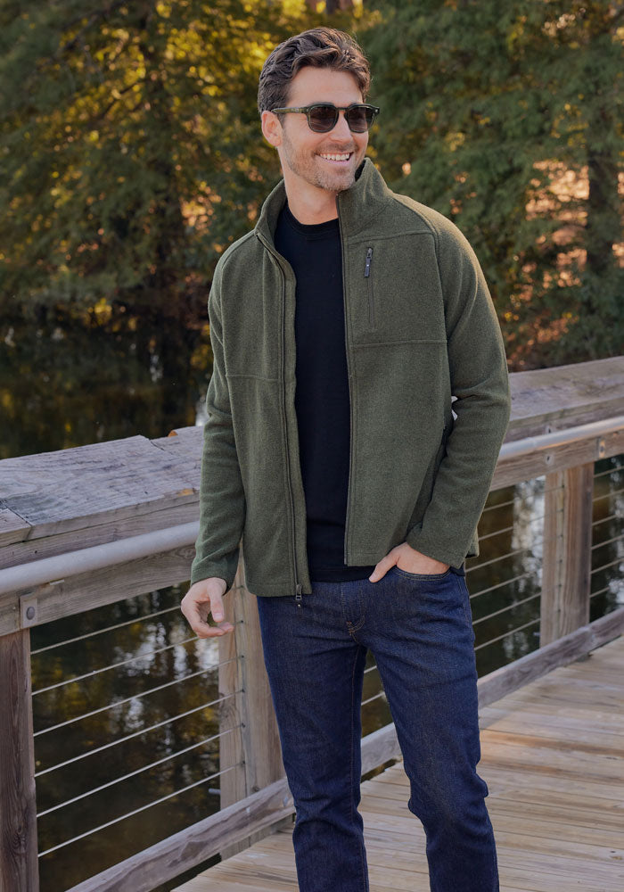 A man wearing sunglasses, a black shirt, blue jeans, and the Woolx Fairbanks Jacket in Dark Navy stands smiling on a wooden bridge with trees and water in the background.