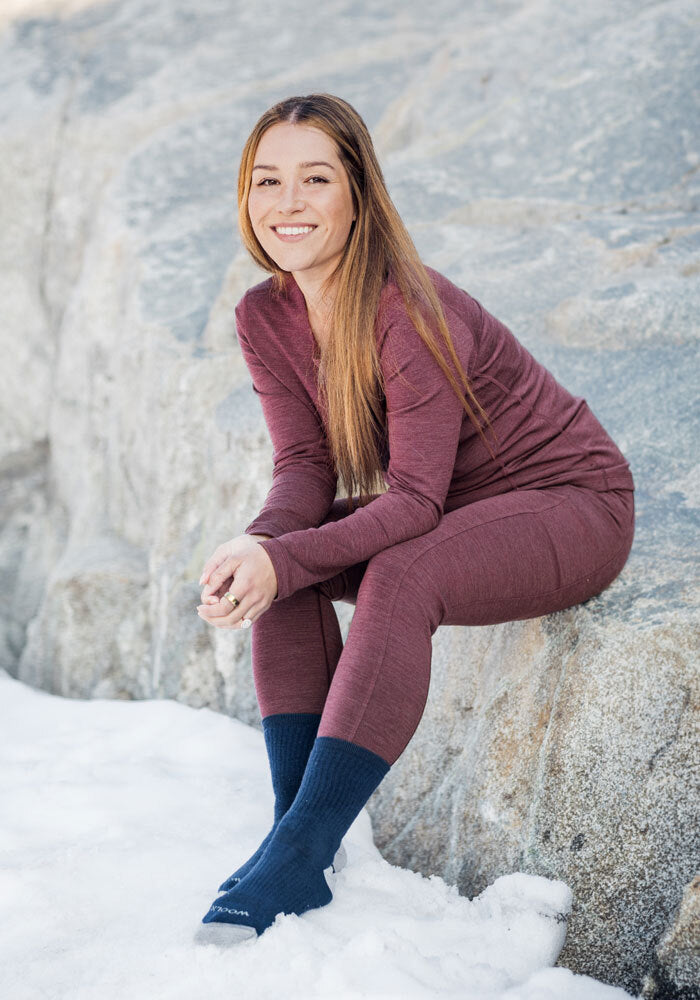 A woman with long brown hair, wearing a maroon top and Woolx Frost Duralite® Leggings in black, sits barefoot except for dark socks on a rock by the snow and smiles at the camera.