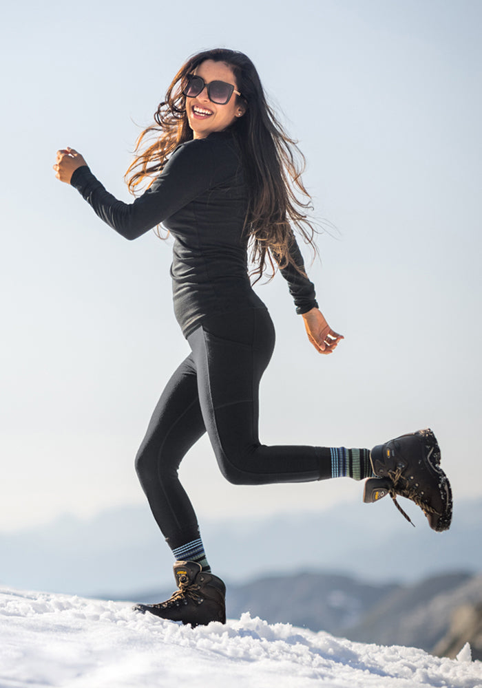 A woman wearing sunglasses, Woolx Frost Petite Duralite® Leggings in black, and hiking boots joyfully runs on snow with mountain scenery in the background.