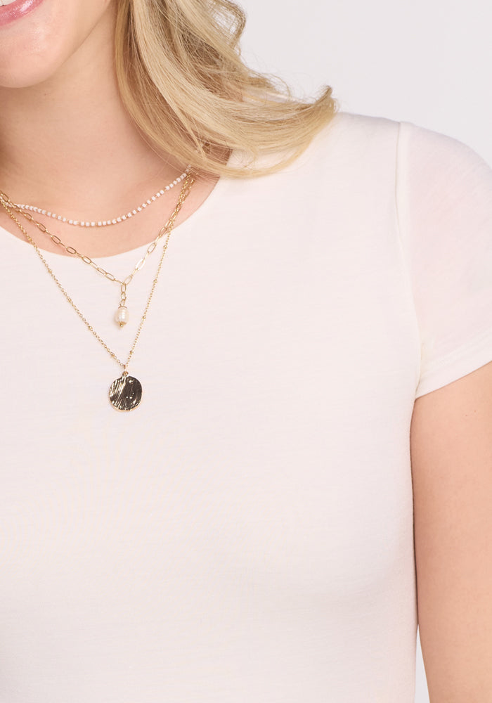 A close-up shows a woman in the Woolx Gracie Bodysuit - Vanilla, prized for its temperature-regulating comfort, paired with layered gold necklaces featuring a round pendant and small pearl. Only her lower face and upper torso are visible.