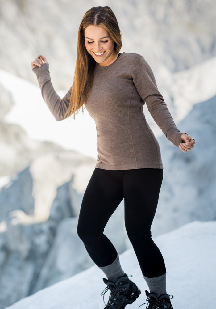 A woman with long brown hair wears the Woolx Hannah Long Sleeve Top in Simply Taupe, black leggings as a thermal base layer, gray socks, and black boots. She smiles while standing on snow with a blurred rocky background.
