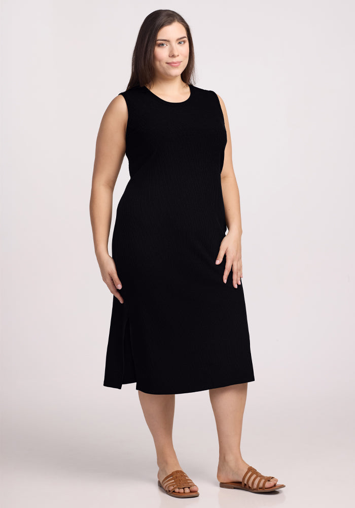 A person stands against a plain background in an Iris Ribbed Dress - Black from Woolx, paired with brown sandals. Their hair is straight and loose, and they have a relaxed expression. | Juliana is 5'10", wearing a size XL