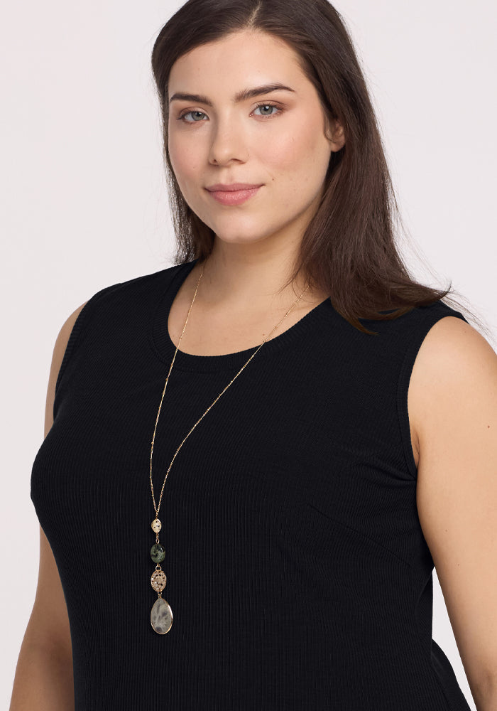 A person with long dark hair wears the Iris Ribbed Dress - Black from Woolx, posing against a plain background. They have an understated smile and a long necklace with decorative pendants.