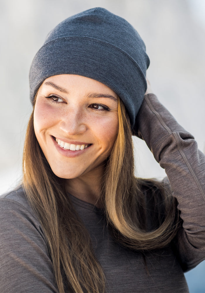 A woman smiles while looking to the side, gently touching her Woolx Journey Hat in Cinder Melange. She wears a long-sleeve brown top, with a softly blurred background behind her.