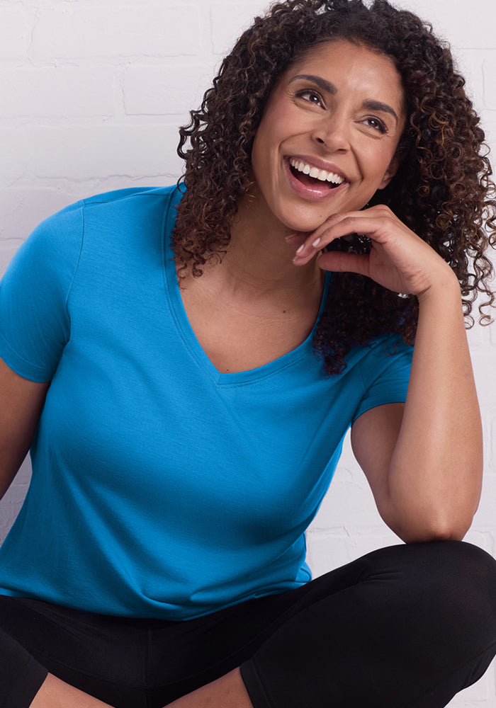 Smiling woman in Woolx Junie V Neck • Final Sale - Wave Rider, relaxed fit, blue tee against a white brick wall.