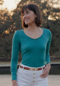 Smiling woman in Woolx Layla V Neck Top in Surfside stands outdoors, wearing white pants with trees behind her.