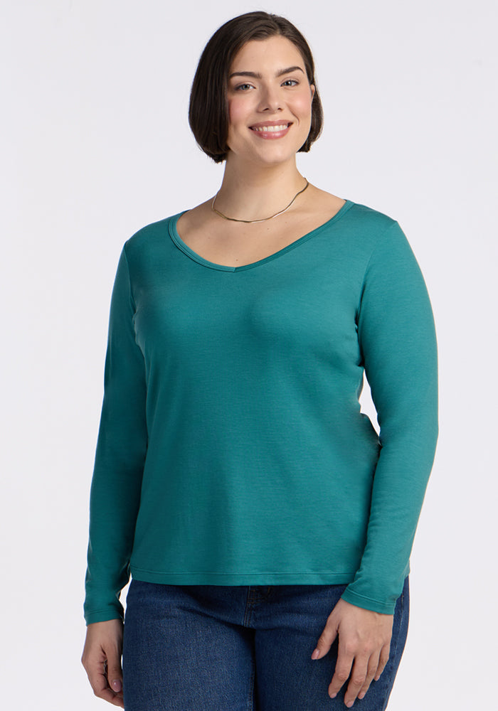 Smiling woman in Woolx Layla V Neck Top - Surfside and jeans stands against a plain white background.