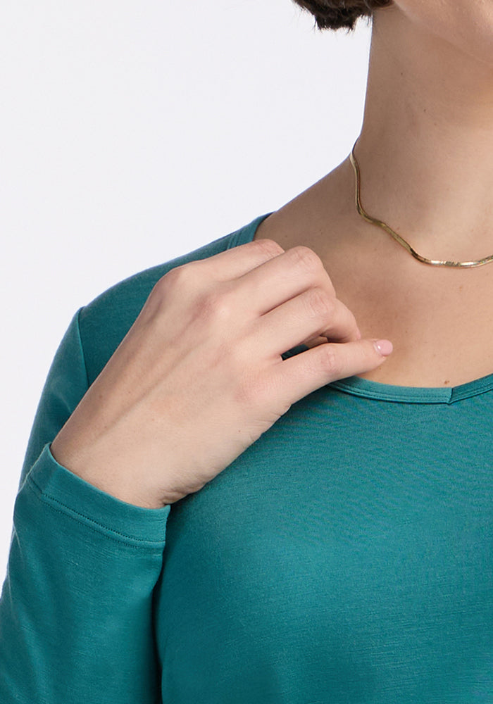 Close-up of a person wearing the Woolx Layla V Neck Top in Surfside and a gold necklace, hand on collarbone.