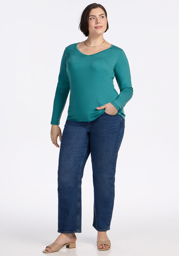 Smiling woman in a Woolx Layla V Neck Top • Final Sale - Surfside and blue jeans, hand in pocket against a white background.