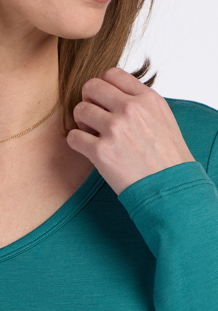 A person in the Woolx Layla V Neck Top in Surfside touches their neck, showing a gold chain and part of their face.