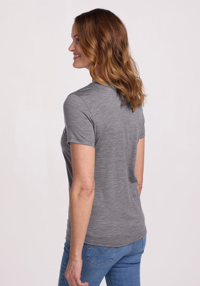 A woman with shoulder-length brown hair smiles, wearing a Woolx Liza Crew Neck Tee in Graphite Heather and blue jeans. She stands slightly turned to the side, hands at her sides, against a plain white background.