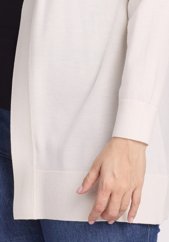 Close-up of a person in the Woolx Macie Sweater - Dewdrop Pearl with ribbed cuffs and hem, wearing blue jeans. Their left hand rests by their side against a neutral background.