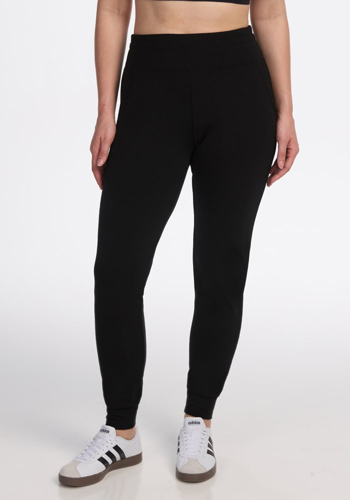 Model wears Woolx Madden Joggers in black, paired with black and white Adidas sneakers and a black crop top, standing against a plain white background. Image shows figure from shoulders down. | Ariana is 5'11", wearing a size S