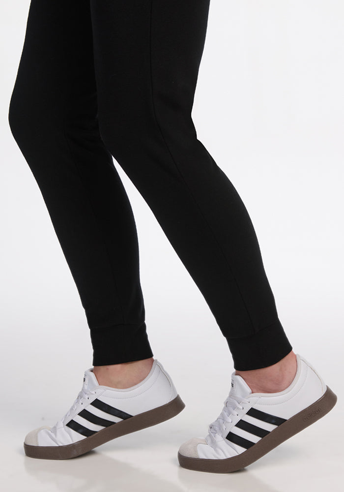 Someone wearing Woolx Madden Petite Joggers in black stands on one leg with the other slightly raised behind, paired with white sneakers featuring black stripes. The background is plain white.
