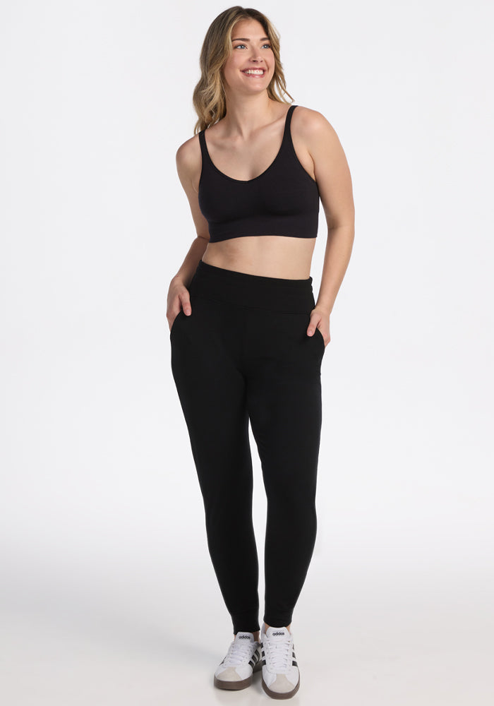 A woman smiles with her hands in the pockets of her black Woolx Madden Petite Joggers, paired with a black sports bra and white sneakers, standing against a plain white background.