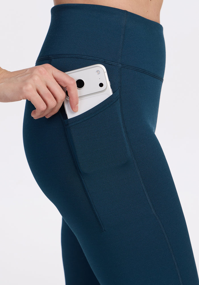 A person in Woolx Merino Flex® McKenna Petite leggings in Majolica Blue slips a white smartphone into the side pocket of the high-waisted athleisure leggings.