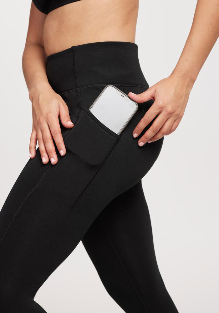 A person wearing Woolx Merino Flex® McKenna Petite leggings places a smartphone into a side pocket, with only their lower torso and hands visible against a plain background.