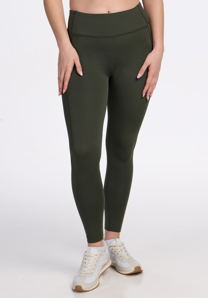 Model wears Woolx Merino Flex® McKenna Pocket Leggings in Everleaf, paired with white sneakers, standing against a plain white background. Their hands rest on their thighs; upper body is out of frame. | Ariana is 5'11", wearing a size S