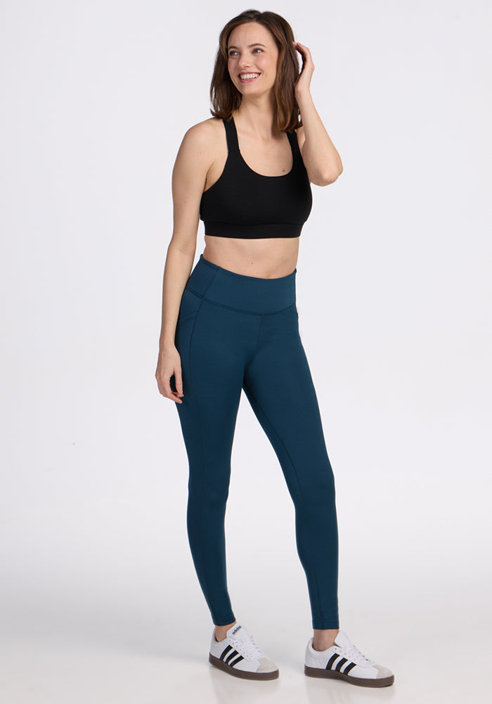 A woman smiles, wearing a black sports bra, Woolx Merino Flex® McKenna Pocket Leggings in Majolica Blue, and white sneakers with black stripes, set against a plain white background.