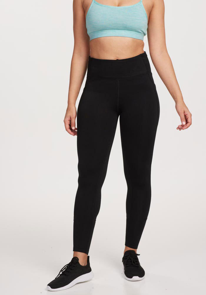 A woman stands against a white background wearing Woolx Merino Flex® McKenna Tall high-waisted black leggings with side pockets and a light blue sports bra. Her arms are relaxed at her sides, and her head is not visible.