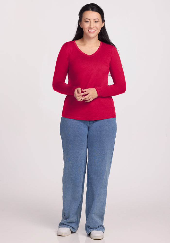 Woman smiling in a Woolx Millie Long Sleeve in Poppy Spark, blue jeans, and white shoes against a plain background.