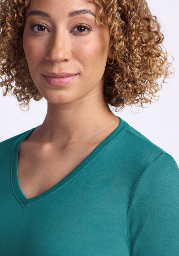 A woman with curly hair smiles softly, wearing the Woolx Millie Long Sleeve in Surfside, shown from the shoulders up.