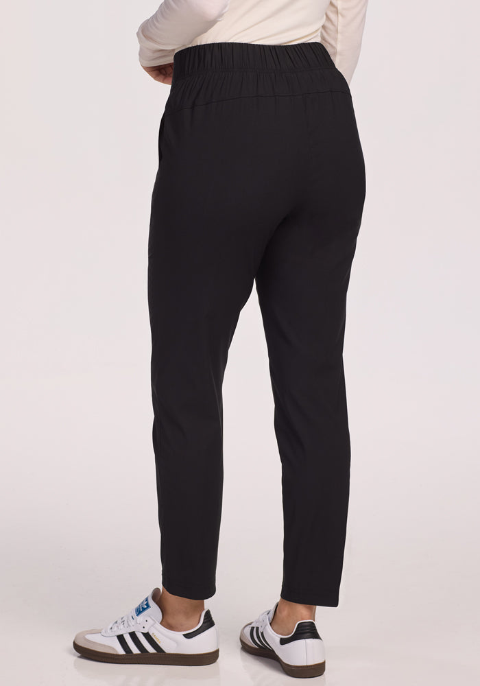 A person is standing with their back to the camera against a plain light background, wearing Woolx Nicole Pants in black and white sneakers with black details.