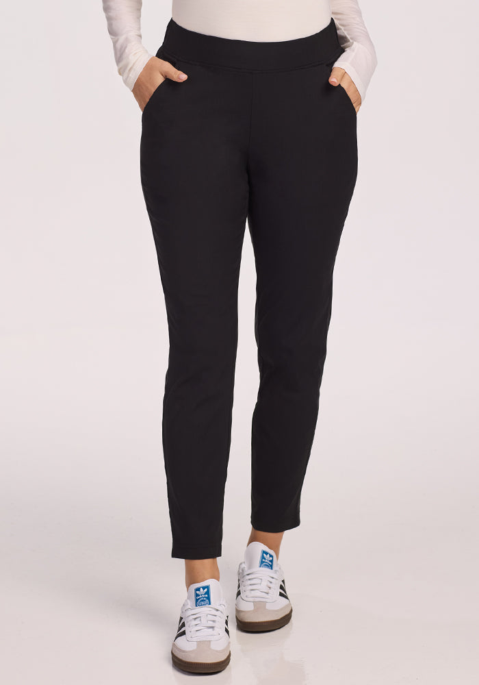 A person wearing Woolx Nicole Pants in black with hands in pockets, a light long-sleeve top, and white sneakers featuring blue logos, standing against a plain white background.