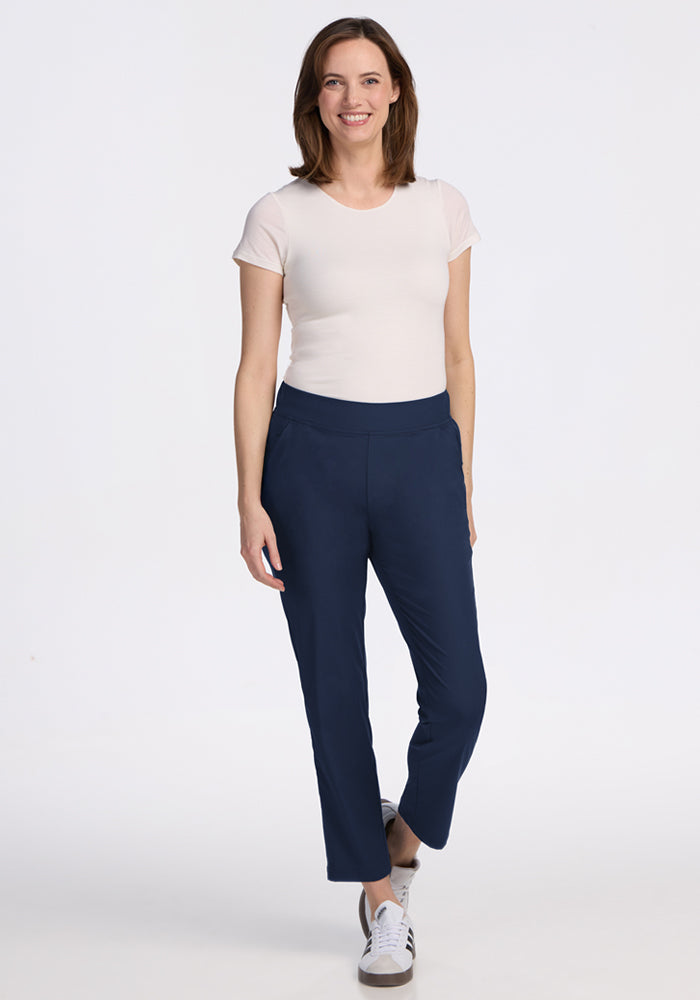 A woman with shoulder-length brown hair smiles against a white background, wearing a short-sleeve white t-shirt, Woolx Nicole Ankle Pants in Deep Navy with a tapered-leg fit, and white sneakers.