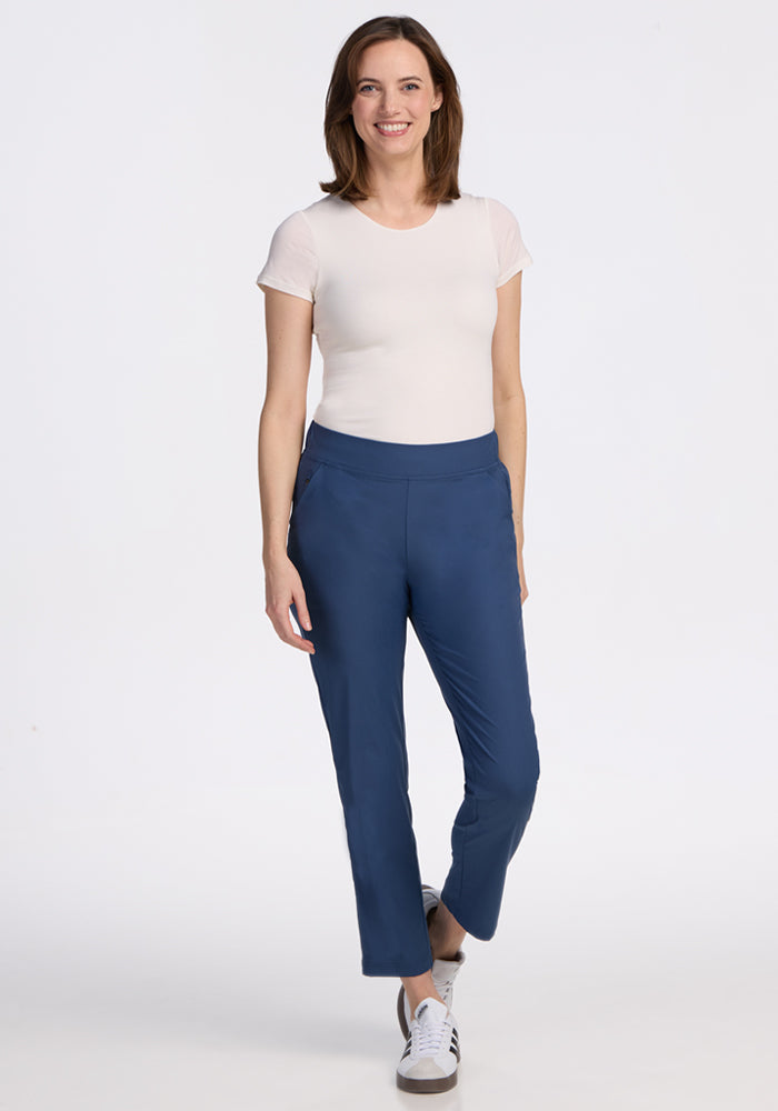 A woman with shoulder-length brown hair smiles while wearing a white short-sleeve top, Woolx Nicole Ankle Pants in Vintage Indigo, and white sneakers. She stands against a plain white background.